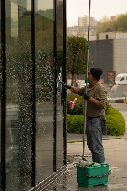 A male window cleaner wearing a black beanie, grey long-sleeve shirt, and blue jeans is using a squeegee and an extension pole to clean large glass panels of a modern shopfront during daylight. The glass is wet with water and soap suds, reflecting the surrounding environment, with visible streaks and soap residue. He is standing on a paved sidewalk beside a green bucket containing cleaning tools, and is in the process of achieving a streak-free shine on the glass surface. Behind him, there are bushes, trees, and a parking lot with cars, indicating an outdoor commercial area. The scene is illuminated by natural light, emphasizing the clarity and cleanliness of the glass surfaces, with clean and polished windows as part of a professional window cleaning service by Swiss Cottage Cleaner.