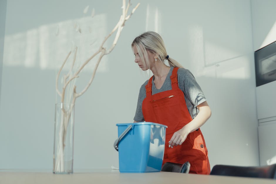 A woman wearing a gray t-shirt and red apron stands in a modern kitchen, engaging in surface cleaning. She holds a blue plastic bucket with a metal handle, positioned on a light-colored countertop. The background features a white wall with natural light illuminating the scene, highlighting the shiny ceramic tiles and sleek stainless steel appliances. The woman's focus appears to be on the cleaning process, emphasizing hygiene and maintenance in domestic cleaning. Swiss Cottage Cleaner specializes in deep cleaning and sanitisation, ensuring kitchens are spotless and hygienic.