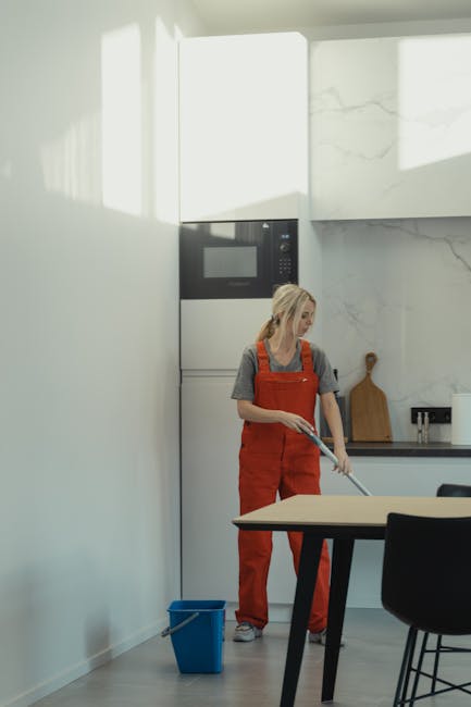 A woman wearing red overalls and a gray shirt is performing surface cleaning in a modern kitchen, using a mop or cloth to clean the floor near a blue cleaning bucket. The kitchen features a white wall with a built-in microwave oven, a black countertop with a wooden cutting board, and black dining chairs around a light-colored table. The surfaces, including the microwave and countertops, are clean and well-maintained, with natural light illuminating the space. This scene exemplifies domestic cleaning and surface hygiene, reflecting the services offered by Swiss Cottage Cleaner as part of their flat cleaning tips for Swiss Cottage Finchley Road area.